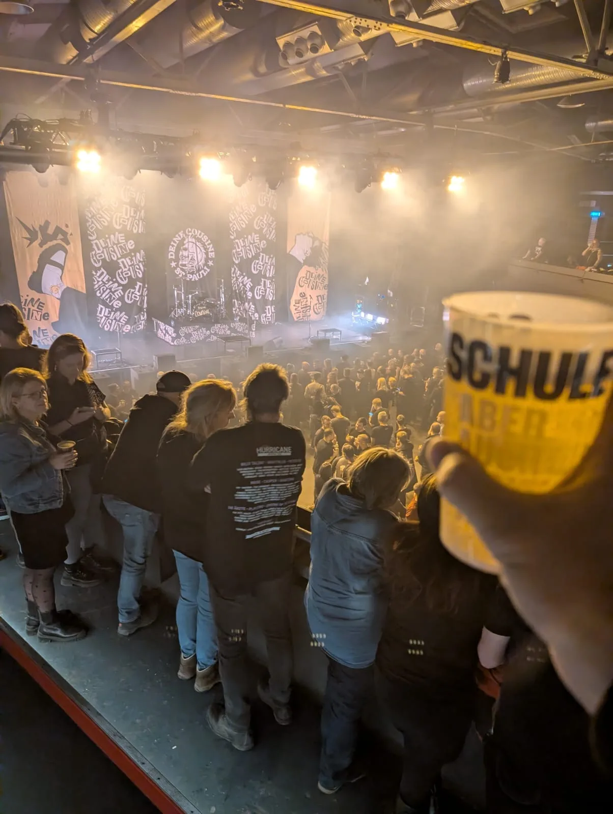 Konzertpublikum von hinten, Banner über der Bühne, ein Becher im Vordergrund