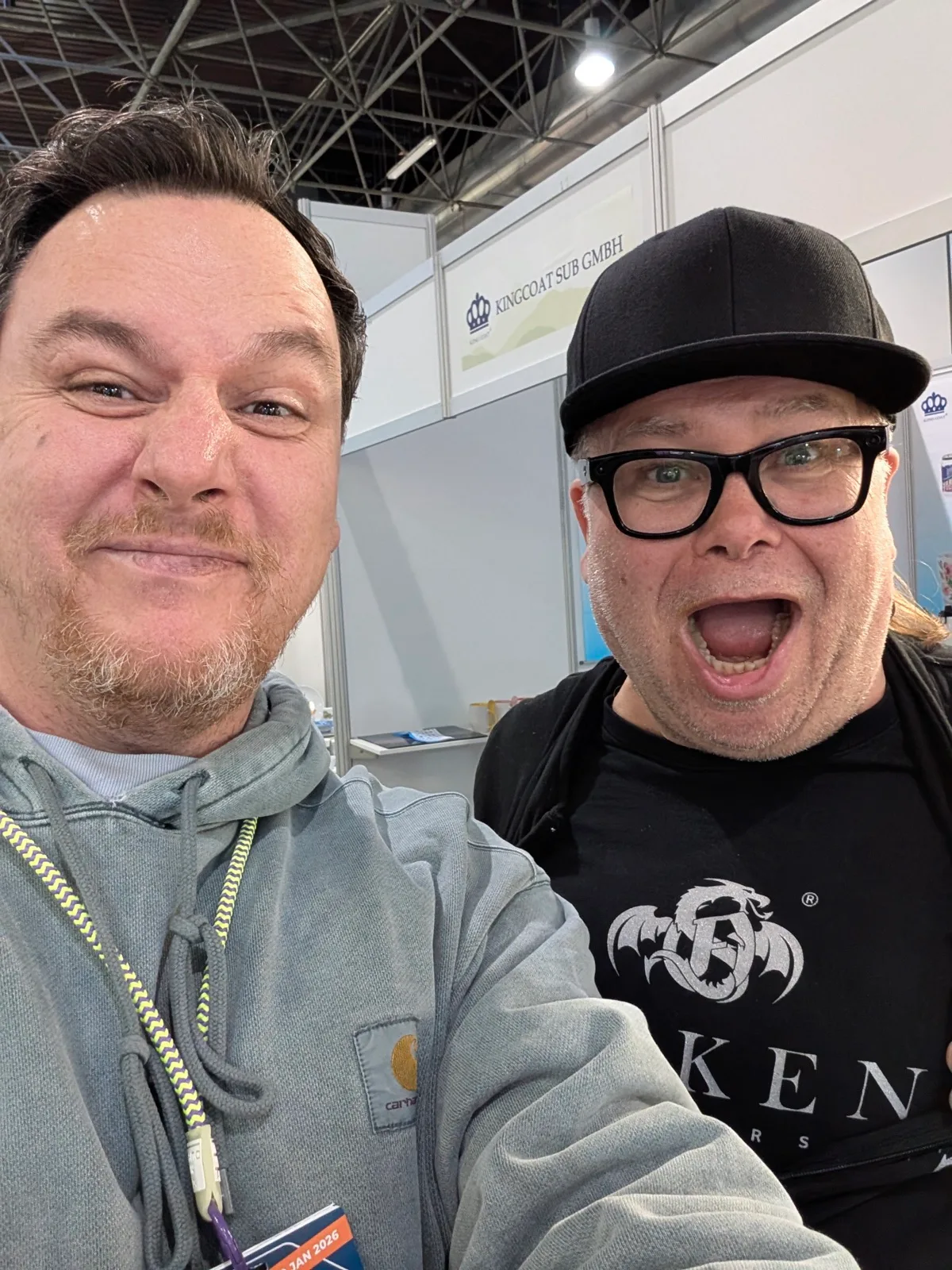 Selfie von zwei Männern auf einer Messe, dahinter ein Ausstellungsstand