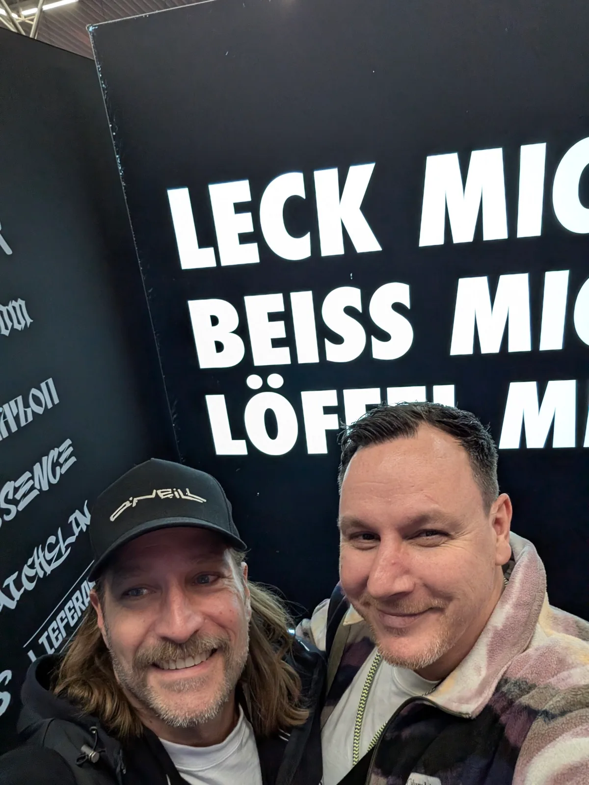 Zwei Männer vor einer schwarzen Wand mit großen weißen Schriftzügen