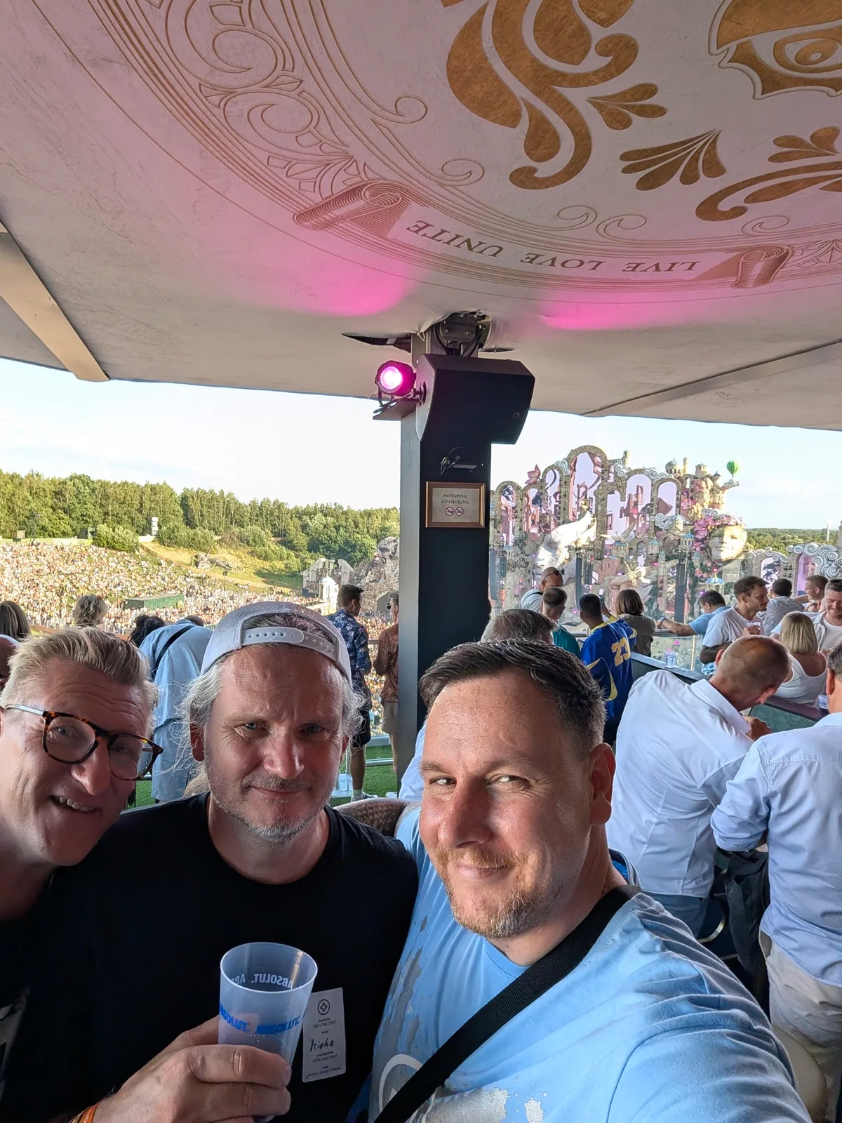 Selfie von drei Männern auf einer Tribüne, dahinter eine große Festivalbühne