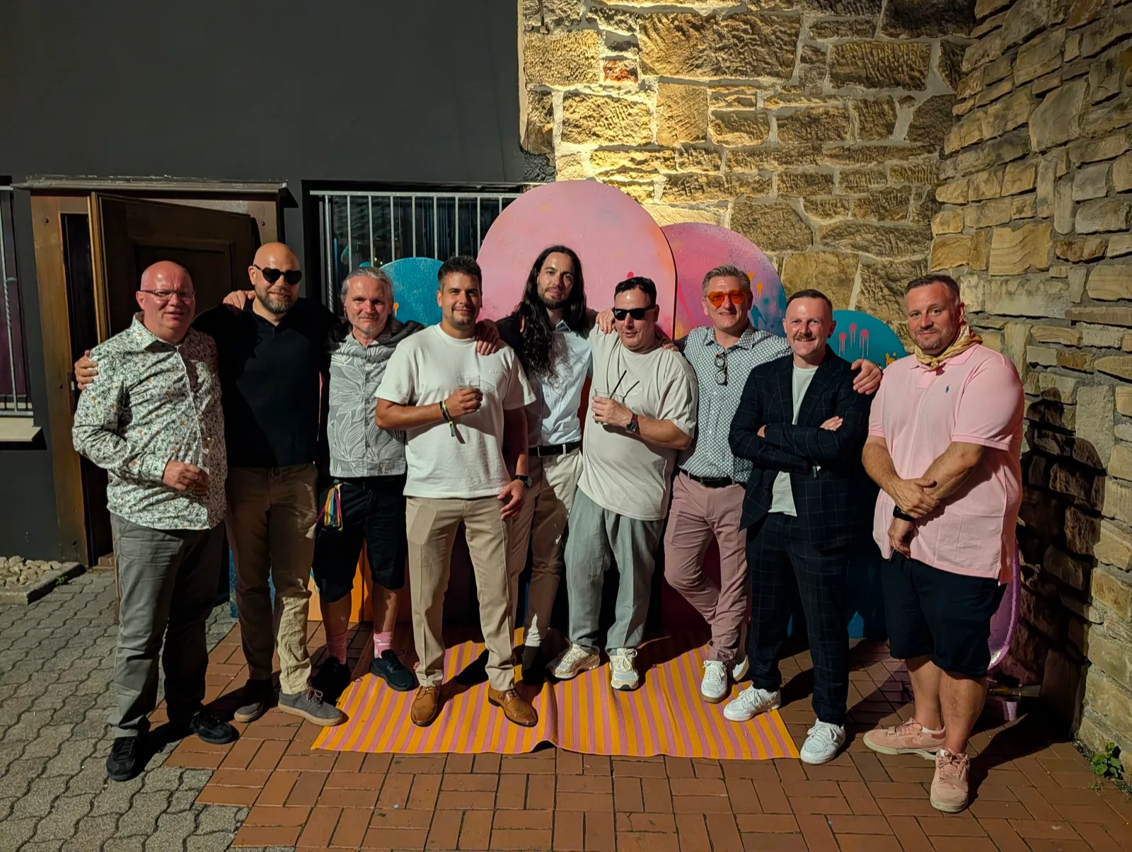 Gruppenfoto von neun Männern vor einer Steinmauer, sommerlich gekleidet