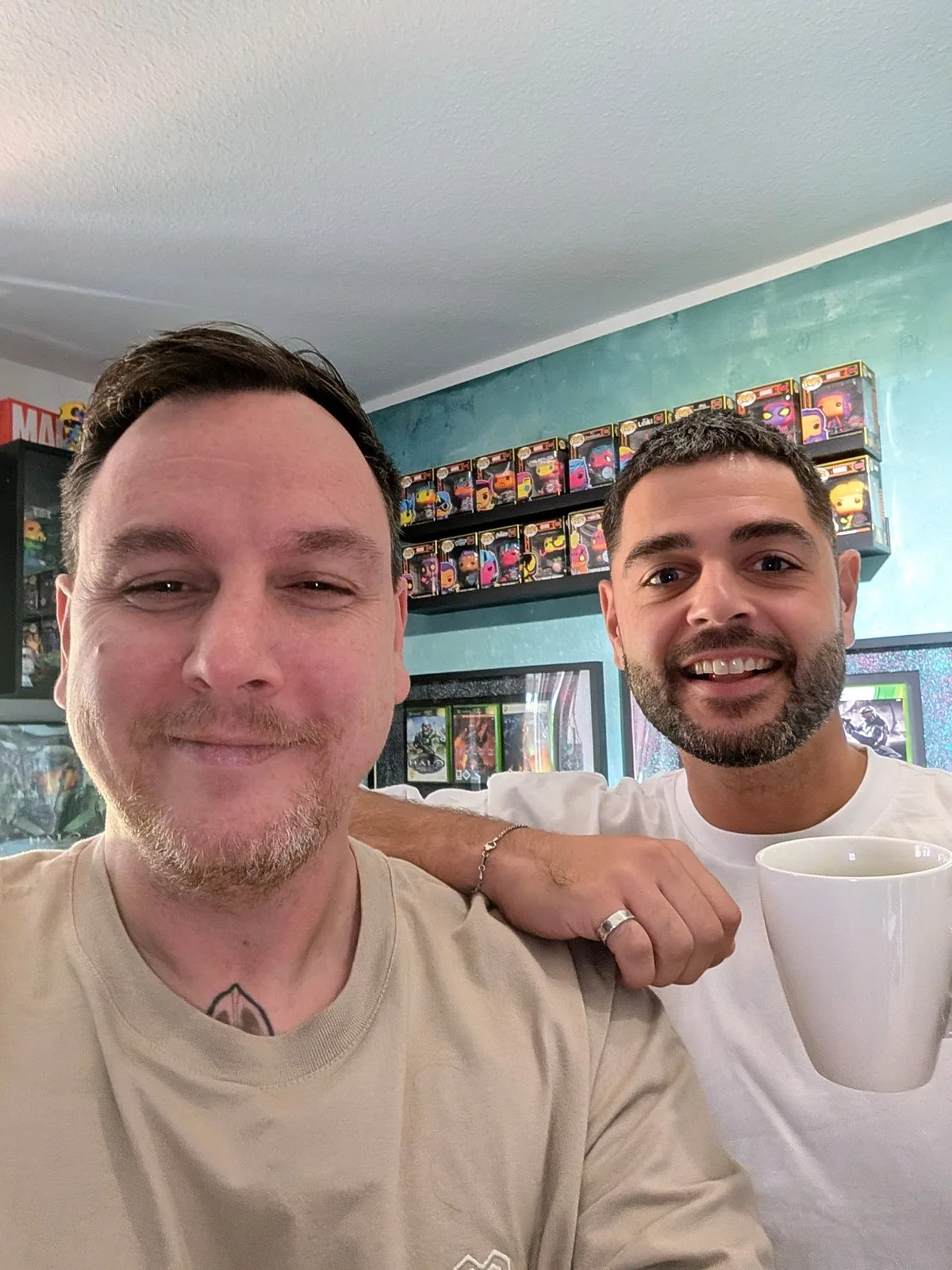 Selfie von zwei Männern vor Wandregalen mit Sammelfiguren, einer hält einen Becher