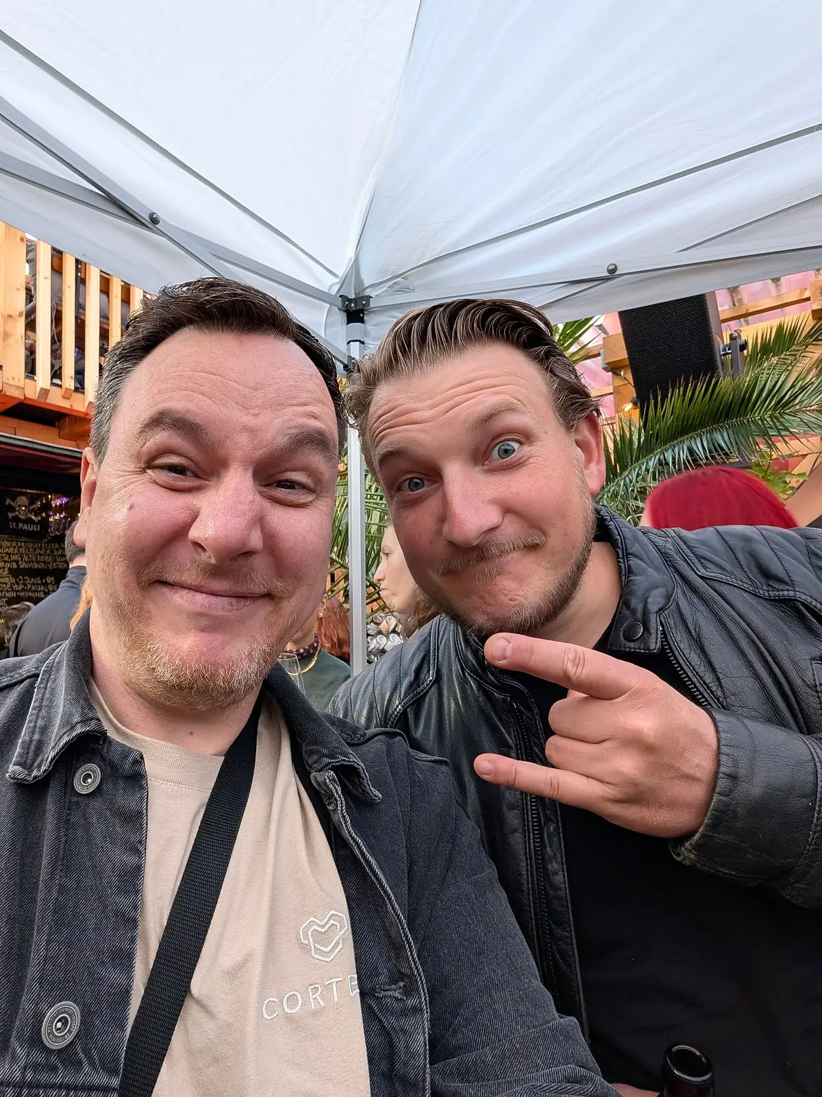 Selfie of two men under a white gazebo, one in a shirt with a heart logo, the other pointing at him