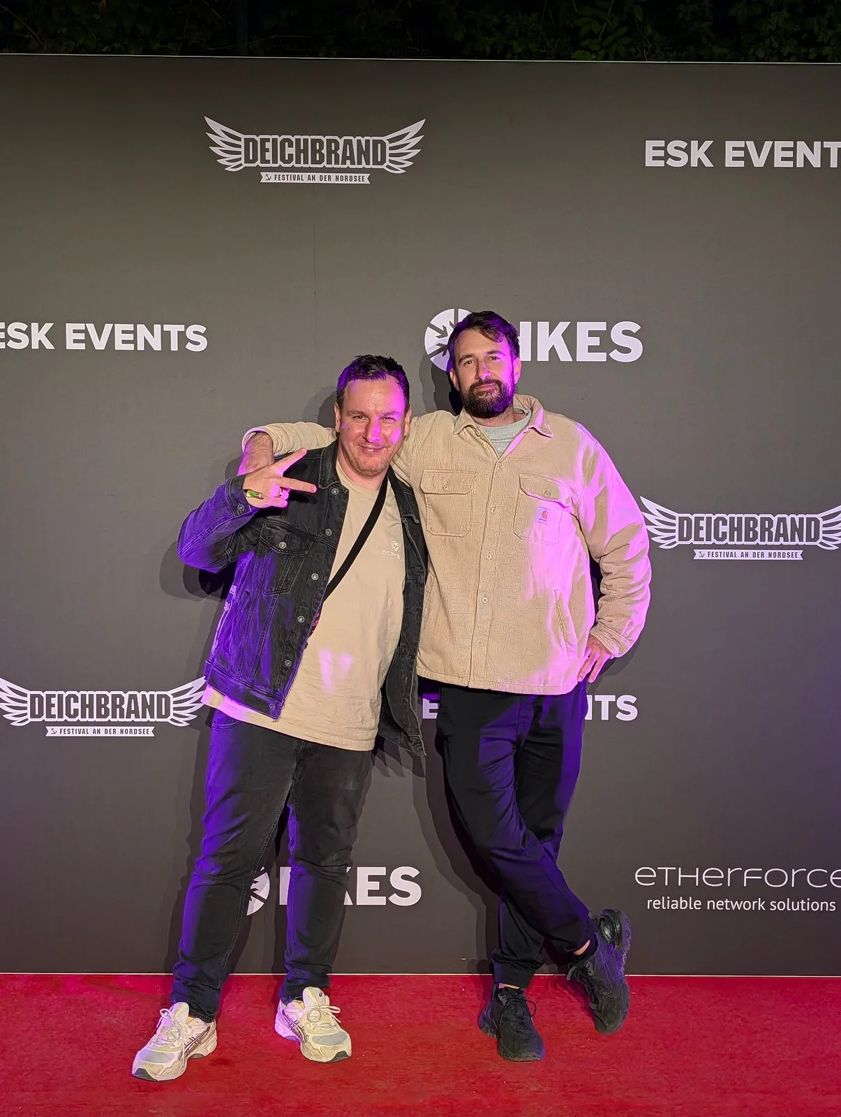 Two men on a red carpet in front of a logo backdrop, one making a victory sign
