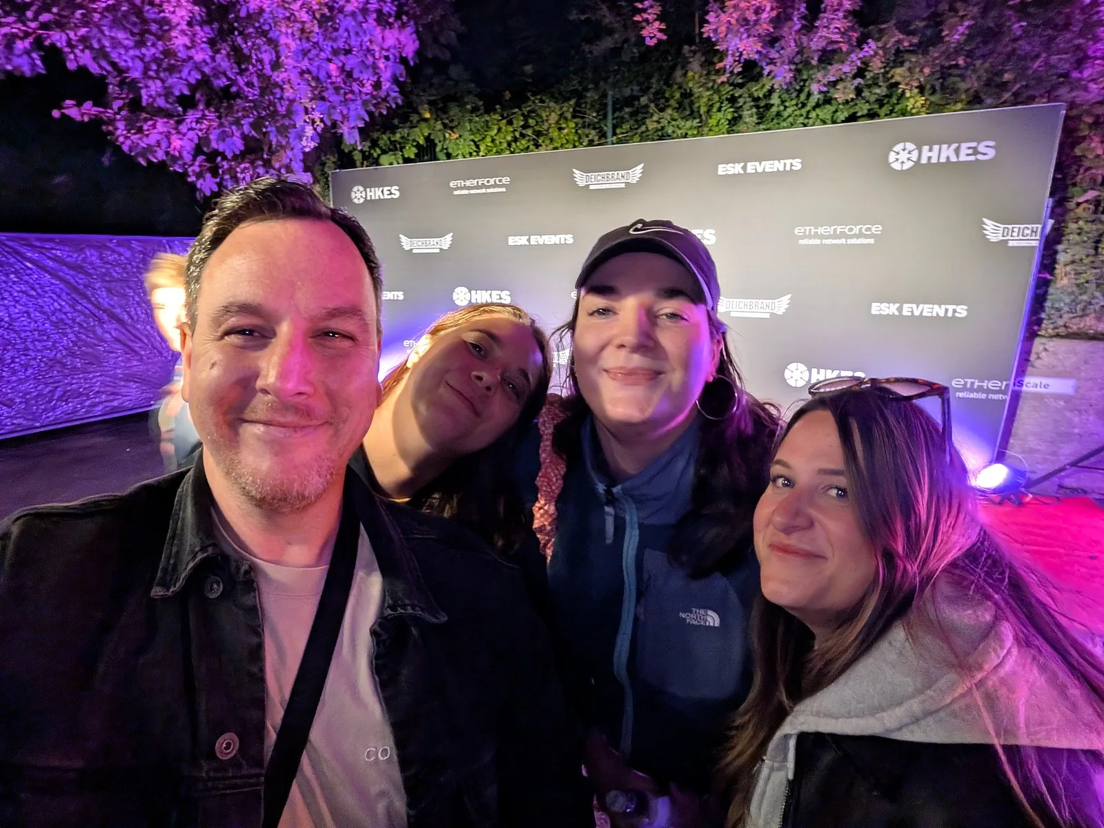 Selfie of a man with three women in the evening in front of a logo backdrop, trees lit in purple