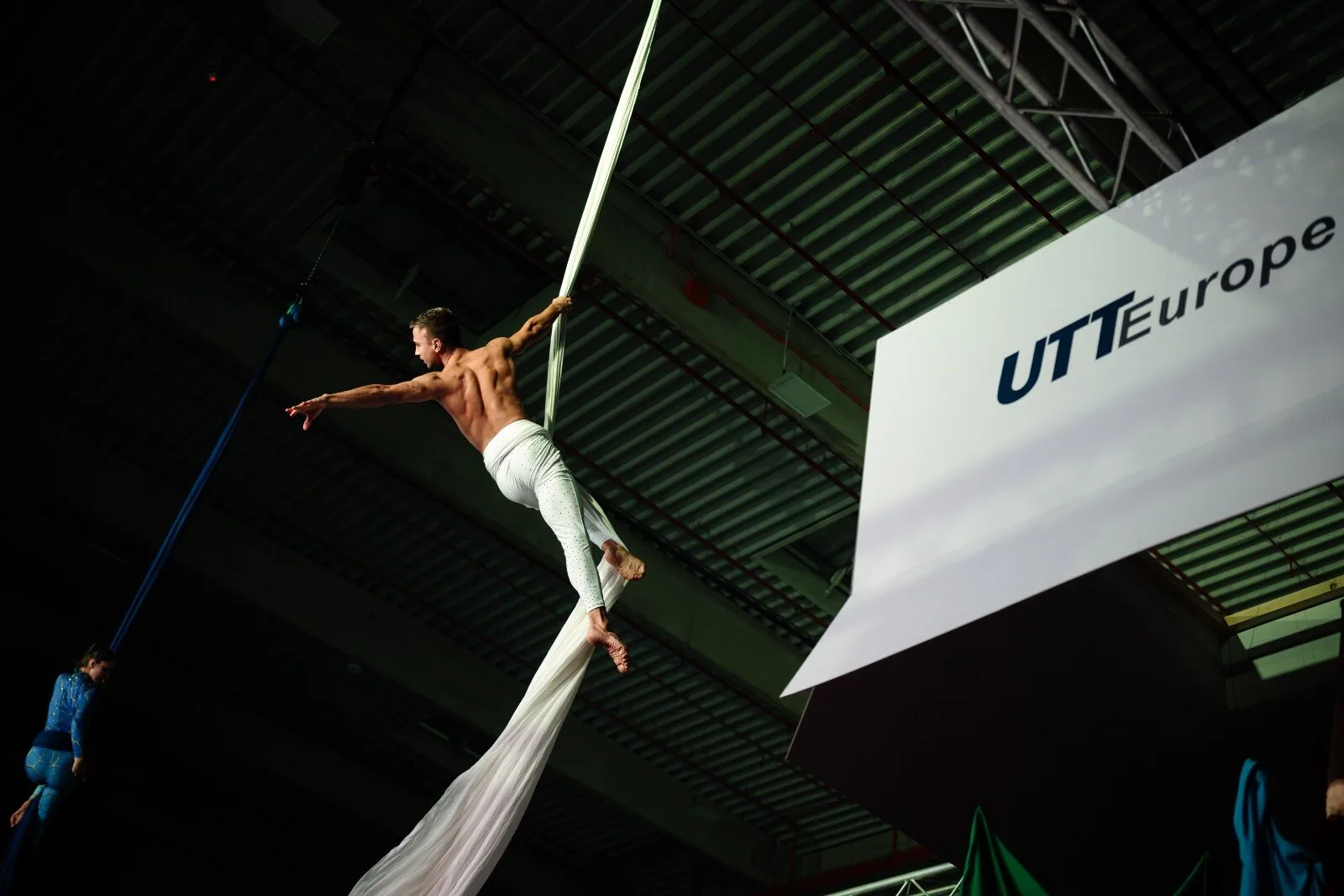 Acrobat on white silks under the hall ceiling, next to a sign reading UTT Europe