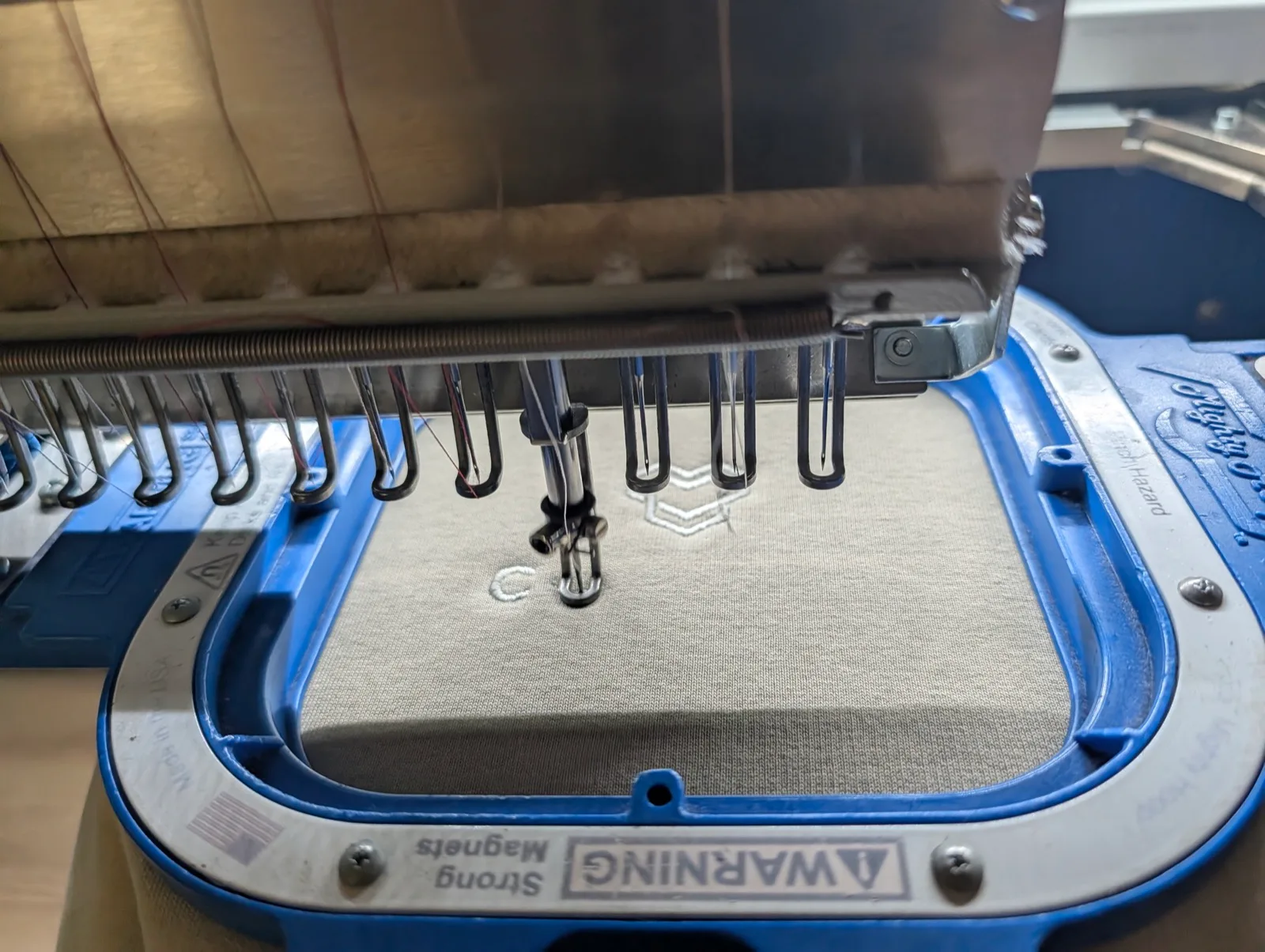 Nadel der Stickmaschine über hellem Stoff im blauen Magnetrahmen, das Signet ist halb gestickt